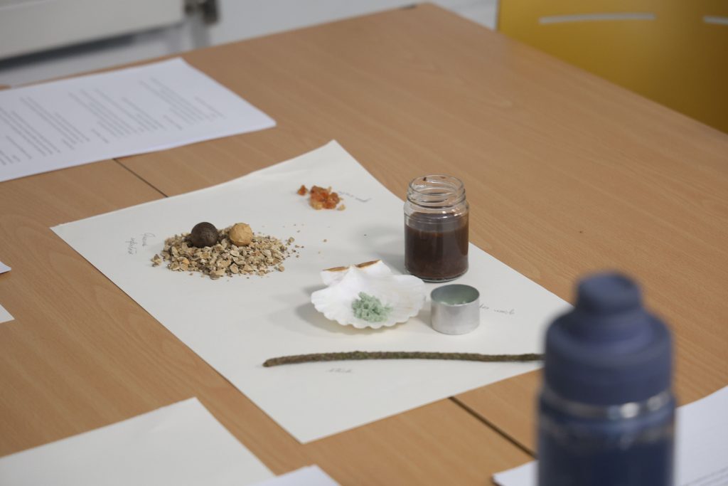 A picture of ink-making materials and a bottle of ink presented on a sheet of laid paper, with iron oxide in a cockle shell